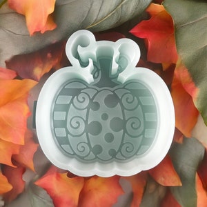 May include: A light blue pumpkin-shaped mold with decorative swirls, stripes, and dots. The mold has a stem and is surrounded by colorful autumn leaves. This item is likely used for crafting or baking.