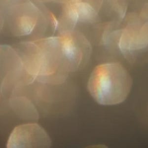 May include: Abstract image with a soft focus effect. The image features out-of-focus, glowing, hexagonal shapes in shades of gold and brown. Some shapes have a rainbow-like iridescence. The overall effect is warm and ethereal.