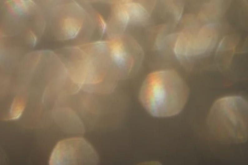 May include: Abstract image with a soft focus effect. The image features out-of-focus, glowing, hexagonal shapes in shades of gold and brown. Some shapes have a rainbow-like iridescence. The overall effect is warm and ethereal.