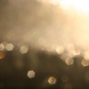 May include: Abstract image with a soft focus effect. The background is a gradient of warm, golden tones, with a bright, blurred light source at the top. Scattered throughout are hexagonal bokeh shapes in shades of gold and brown.