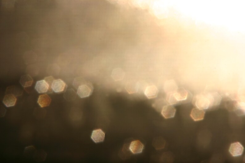 May include: Abstract image with a soft focus effect. The background is a gradient of warm, golden tones, with a bright, blurred light source at the top. Scattered throughout are hexagonal bokeh shapes in shades of gold and brown.