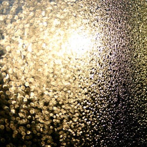 Könnte beinhalten: Nahaufnahme einer strukturierten Oberfläche mit einem verschwommenen Hintergrund aus durchscheinendem Licht. Die Oberfläche ist mit kleinen, runden, goldfarbenen Tropfen bedeckt.