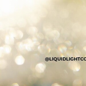K&ouml;nnte beinhalten: Abstraktes Bild mit Weichzeichner-Effekt, das einen Bokeh-Effekt aus schimmernden, unscharfen Lichtern in Gold- und Wei&szlig;t&ouml;nen zeigt. Der Text "@LIQUIDLIGHTCODES" ist in Schwarz dargestellt.