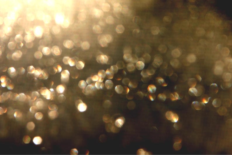 May include: An abstract image with a blurred focus, showing many golden, circular bokeh lights against a dark background. The lights vary in size and intensity, creating a shimmering, ethereal effect. The overall impression is one of warmth and radiance.