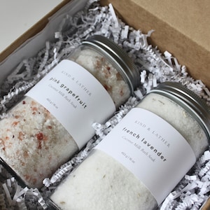 May include: Two glass jars filled with bath salts in a brown cardboard box. The jars have white labels with black text that reads "KIND & LATHER" and the scent of the bath salts. The first jar is labelled "pink grapefruit Coconut Milk Bath Soak 403 g/14.2 oz". The second jar is labelled "french lavender Coconut Milk Bath Soak 403 g/14.2 oz".