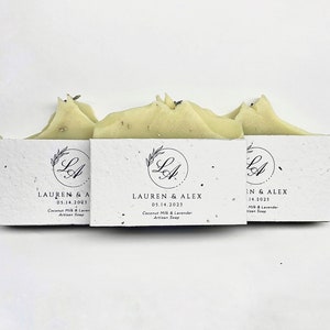May include: Three bars of handmade soap with a white paper wrapper. The wrapper has the text "LAUREN & ALEX 05.14.2023 Coconut Milk & Lavender Artisan Soap" printed on it.