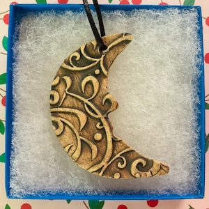 May include: A crescent moon pendant with an intricate, swirling design. The pendant is a warm brown color with darker brown detailing. It hangs from a black cord and is presented in a blue box with white filler.