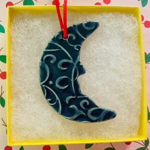 May include: A ceramic crescent moon ornament with a teal glaze and white swirling patterns. The ornament is displayed in a yellow box with white filler and a red ribbon for hanging. The background has a floral pattern.