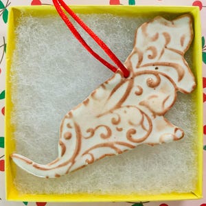 May include: A ceramic dog ornament with brown scrollwork details, presented in a yellow box with white filler and a red hanging string. The ornament has a glossy finish and is likely handmade.