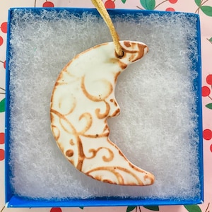 May include: A ceramic crescent moon ornament with a cream-colored base and brown decorative swirls. The ornament is presented in a blue box with white padding and a gold string for hanging. The ornament has a face carved into the moon.