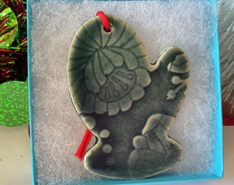 May include: A ceramic mitten-shaped Christmas ornament in a light blue gift box. The ornament is a muted green with a floral design and a red string for hanging. The box is lined with white, fluffy material.