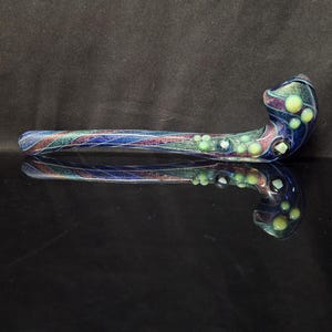 May include: A long, curved glass pipe featuring a deep blue and purple base with iridescent, glittery streaks. The surface is decorated with small, raised, pale green circular glass beads and accents, creating a textured appearance along the stem.