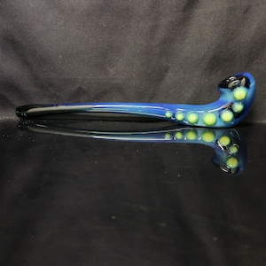 May include: A blue glass pipe with yellow and green polka dots. The pipe has a curved design with a black mouthpiece and a long, slender stem. The glass reflects on the black surface.