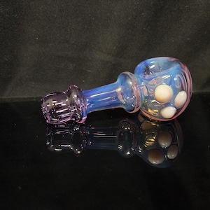 May include: A hand pipe made of iridescent glass featuring a translucent blue and pink color gradient. The bowl section is rounded and decorated with three opaque white circular glass beads arranged in a triangular pattern on the surface.