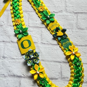 Puede incluir: Un lei de graduación hecho a mano en verde y amarillo, con los colores de la Universidad de Oregón. El lei está adornado con flores de tela y el logotipo de los Oregon Ducks. La cinta está trenzada y tiene un lazo decorativo.