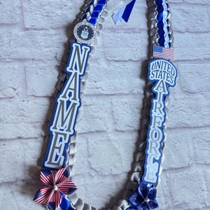 Könnte beinhalten: Eine dekorative Band-Lei aus silbernen und königsblauen Satinbändern. Sie enthält ein kreisförmiges Air-Force-Emblem, eine Flagge der Vereinigten Staaten und vertikale Ausschnitte mit der Aufschrift NAME und UNITED STATES AIR FORCE. Die Basis ist mit Stoffblumen verziert.