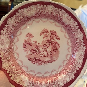 May include: A round ceramic plate with a scalloped edge, featuring a deep red transferware pattern on a cream background. The center depicts a pastoral scene with figures and a dog, surrounded by an ornate border of scrolls and lattice.