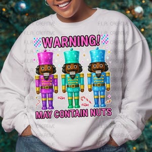May include: White sweatshirt with a graphic of three colorful nutcrackers and the text "WARNING! MAY CONTAIN NUTS." The nutcrackers are in purple, teal, and blue with gold accents. The background is a Christmas tree.