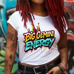 Op de afbeelding: Wit T-shirt met de tekst "Big Gemini Energy" in een kleurrijke graffiti-stijl. Het shirt heeft een kroongrafiek boven de tekst. Het model heeft rood-zwarte dreadlocks en draagt een zwarte riem en jeans.