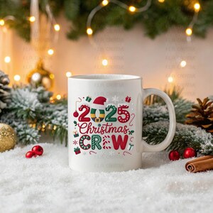 A white mug with a festive Christmas-themed design sits on a snowy surface, surrounded by holiday decorations such as pine cones, ornaments, and a garland with twinkling lights.