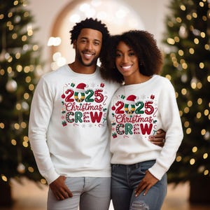 A man and a woman are standing together in front of Christmas trees, both wearing matching white Christmas crew sweaters with the text "2025 Christmas Crew" printed on them.