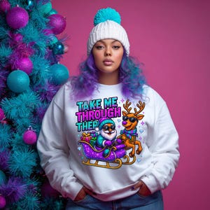 May include: White sweatshirt with a colorful graphic of Santa Claus in a sleigh pulled by a reindeer, both wearing sunglasses. The text "TAKE ME THROUGH THER" is above the graphic. The person is wearing a white beanie with a blue pom-pom.