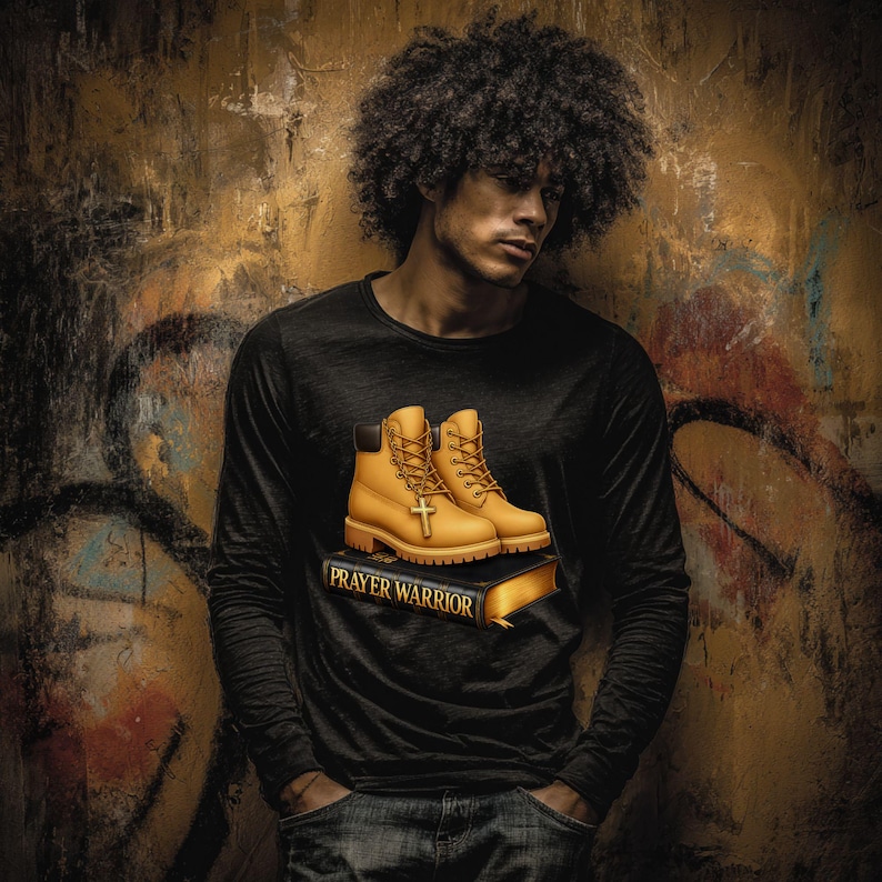 A man with curly hair wearing a black shirt with a graphic of yellow boots and the text "Prayer Warrior" stands in front of a graffiti-covered wall.