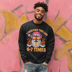 May include: Black sweatshirt with a colorful graphic of Santa Claus wearing sunglasses and a peace sign. The text "I CHECKED THE LIST" and "6-7 TIMES" are displayed in a playful font. The design features hearts and dollar signs.