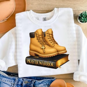 A white t-shirt with a graphic design of yellow boots on top of a black book titled "Prayer Warrior".