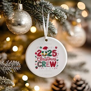 A white ornament with the text "2025 Christmas Crew" is hanging on a Christmas tree, surrounded by other ornaments and lights.