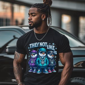 May include: Black t-shirt featuring the text "THEY NOT LIKE US" above a graphic of three snowmen wearing sunglasses, hoodies, and chains. The snowmen are in purple, blue, and black outfits. The shirt is short-sleeved.