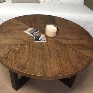 May include: A round wooden coffee table with a chevron pattern top and black metal legs. The table has a dark brown stain and is decorated with two coasters and a candle.