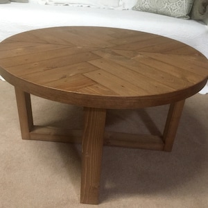 May include: A round wooden coffee table with a herringbone pattern top and a simple, sturdy base. The table is made of natural wood and has a warm brown finish.