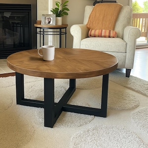 May include: A round coffee table with a light brown wooden top and black metal legs. The table is approximately 36 inches in diameter and sits on a cream-colored rug. A white mug rests on the table's surface.