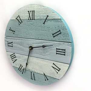 May include: A round wall clock with a light blue and white wooden face. The clock features black Roman numerals and black clock hands. The clock's outer edge is a solid light blue color.