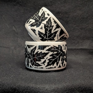 Puede incluir: Dos cuencos de cerámica blanca con un patrón de hojas negras. Un cuenco está apilado sobre el otro. Los cuencos tienen forma cilíndrica con un borde ligeramente ensanchado. El diseño de hojas negras cubre el exterior de los cuencos.