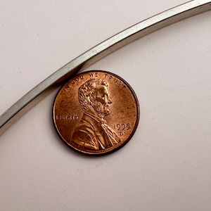 May include: A copper penny from 1995 with the profile of Abraham Lincoln, next to a silver metal band. The penny is a warm brown color, and the band is a shiny silver. The penny is slightly angled.