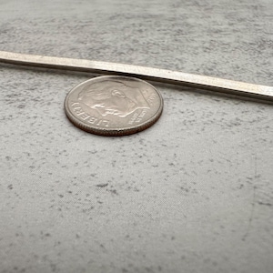 May include: A thin, straight, metallic silver-colored rod positioned horizontally above a United States ten-cent coin, also known as a dime, which features the profile of Franklin D. Roosevelt and the text Liberty and In God We Trust.