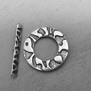 May include: A silver-toned toggle clasp set, featuring a textured bar and a circular ring. The ring has an open center and a patterned design. The clasp is likely for jewelry making or crafts.