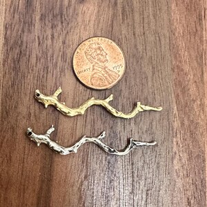 May include: Two metal charms shaped like small, textured tree branches, one in a gold tone and one in a silver tone, positioned below a 1995 United States copper penny for scale. Each charm features a small hole at one end for attachment.