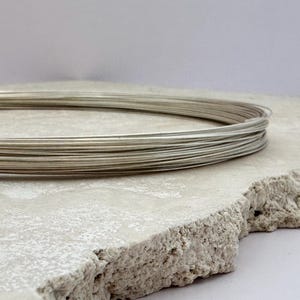 May include: A coil of silver-colored wire sits atop a rough, light-colored surface. The wire is tightly wound, forming a circular shape. The surface has a textured, uneven edge, and the background is a soft, neutral tone.