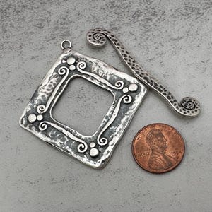 May include: A silver-toned, square pendant with an open center and decorative swirls and dots. A matching bookmark with a spiral end and a penny are also shown. The pendant has a textured, hammered appearance.