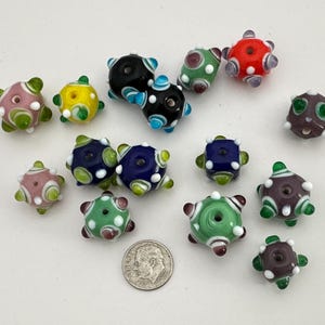 May include: A collection of colorful glass beads in various shapes and sizes. The beads feature a variety of colors, including pink, yellow, blue, green, and red, with white polka dots. A US dime is included for size reference.