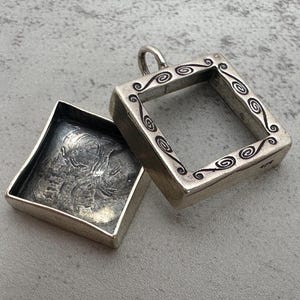 May include: A silver-colored, square pendant with an open frame and decorative scrollwork. The pendant is open, revealing a detailed interior. The lid is open, showing a detailed relief design. The pendant has a loop for a chain.
