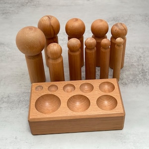 May include: A wooden toy set featuring a variety of wooden peg dolls and a rectangular wooden tray with circular indentations. The peg dolls vary in size and shape, with some having rounded heads. The set is made of light-colored wood.