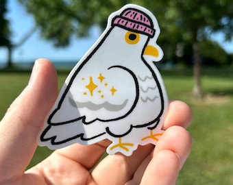May include: A white sticker featuring a cartoon pigeon wearing a pink beanie. The pigeon has yellow eyes and feet, with a black outline and a sparkling design on its side. The sticker is held up against a blurred green background.