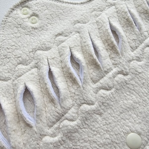 Close-up of a reusable, off-white menstrual pad. The pad has a textured, plush surface with several curved openings and snap closures. The edges are neatly finished with a white trim.