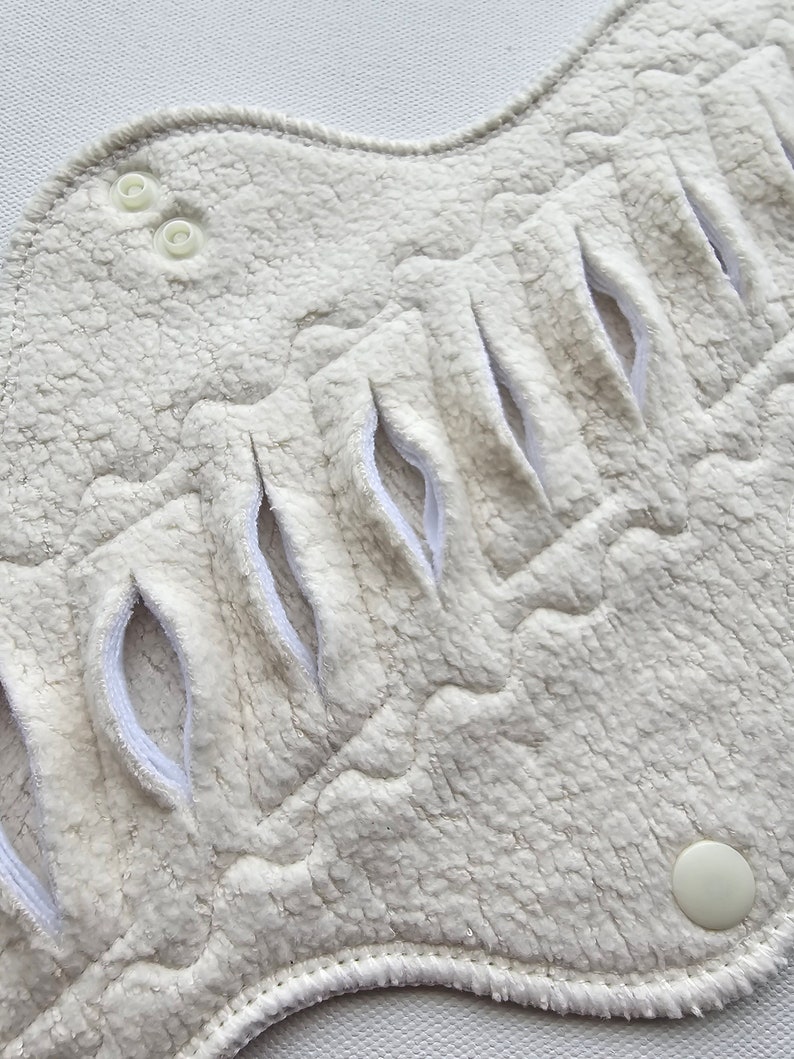 Close-up of a reusable, off-white menstrual pad. The pad has a textured, plush surface with several curved openings and snap closures. The edges are neatly finished with a white trim.