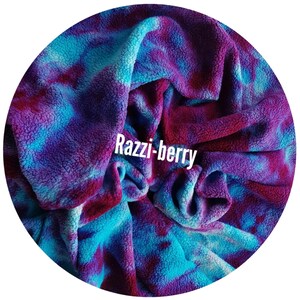 A circular image showing a soft, plush organic cotton sherpa fleece fabric with a tie-dye pattern in shades of blue, purple, and red. The text "Razzi-berry" is displayed in white across the fabric.