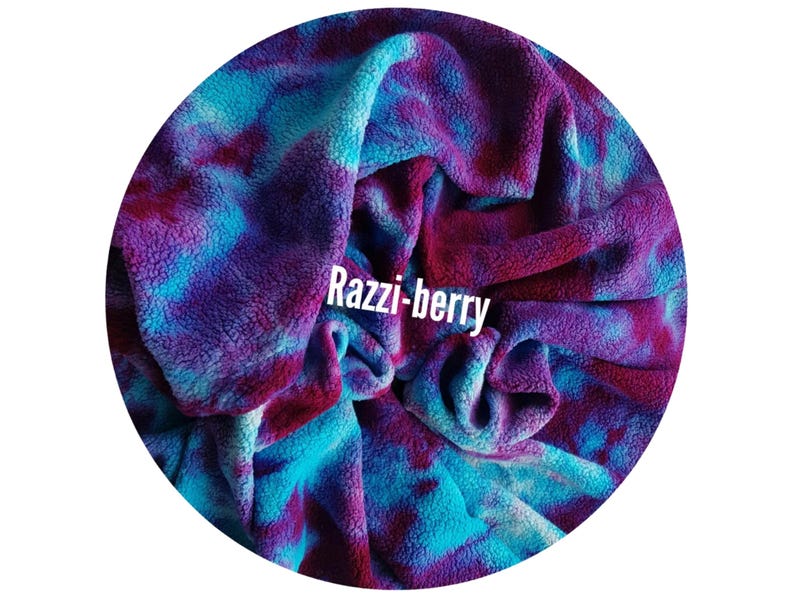 A circular image showing a soft, plush organic cotton sherpa fleece fabric with a tie-dye pattern in shades of blue, purple, and red. The text "Razzi-berry" is displayed in white across the fabric.